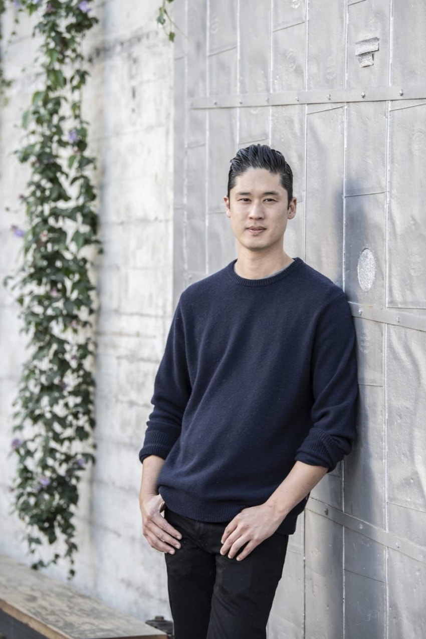 Man in dark sweater leaning against a textured wall, with plants climbing nearby.