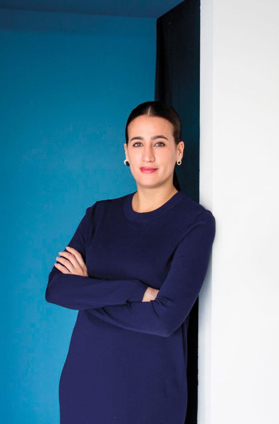 Person in a navy blue outfit, standing with arms crossed, leaning against a black and white wall with a blue background.