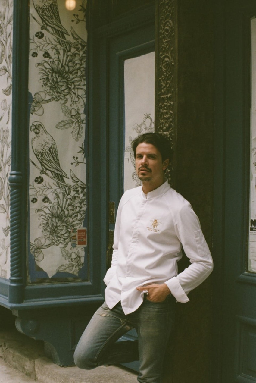 Chef in a white jacket leaning against the entrance of a restaurant with ornate floral and bird design on the window.