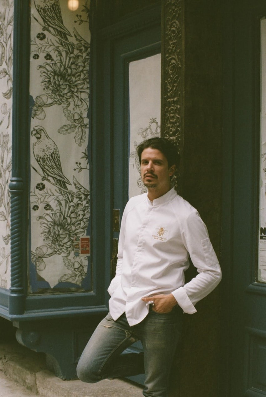 Chef in a white jacket leaning against the entrance of a restaurant with ornate floral and bird design on the window.