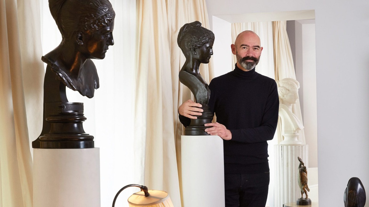 Man standing beside sculptures in a room with curtains and a lamp beside him.