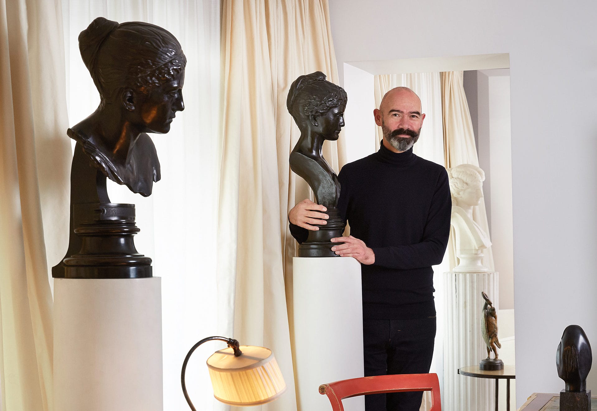 Man standing beside sculptures in a room with curtains and a lamp beside him.