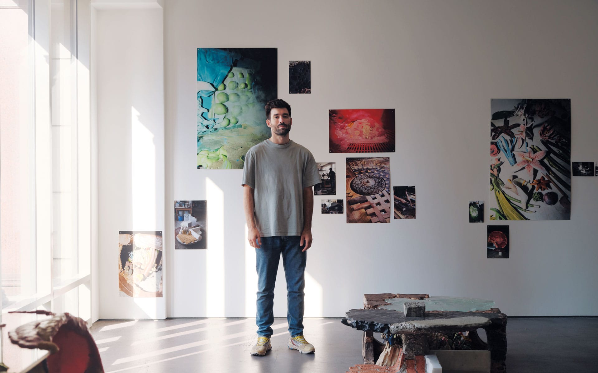 Man stands in an art gallery surrounded by various colorful paintings on a white wall, with sunlight streaming in.