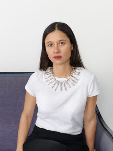 Woman seated wearing a white shirt and a statement necklace against a plain background