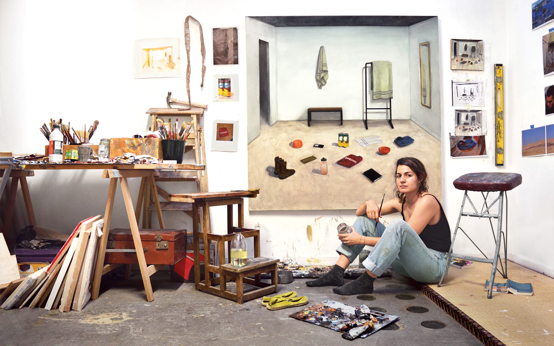 Artist sitting in a studio next to a painting, surrounded by art supplies and tools, in a creative and casual setting.