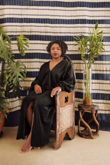 Person sitting on a wooden chair surrounded by plants, in front of a striped cloth backdrop.