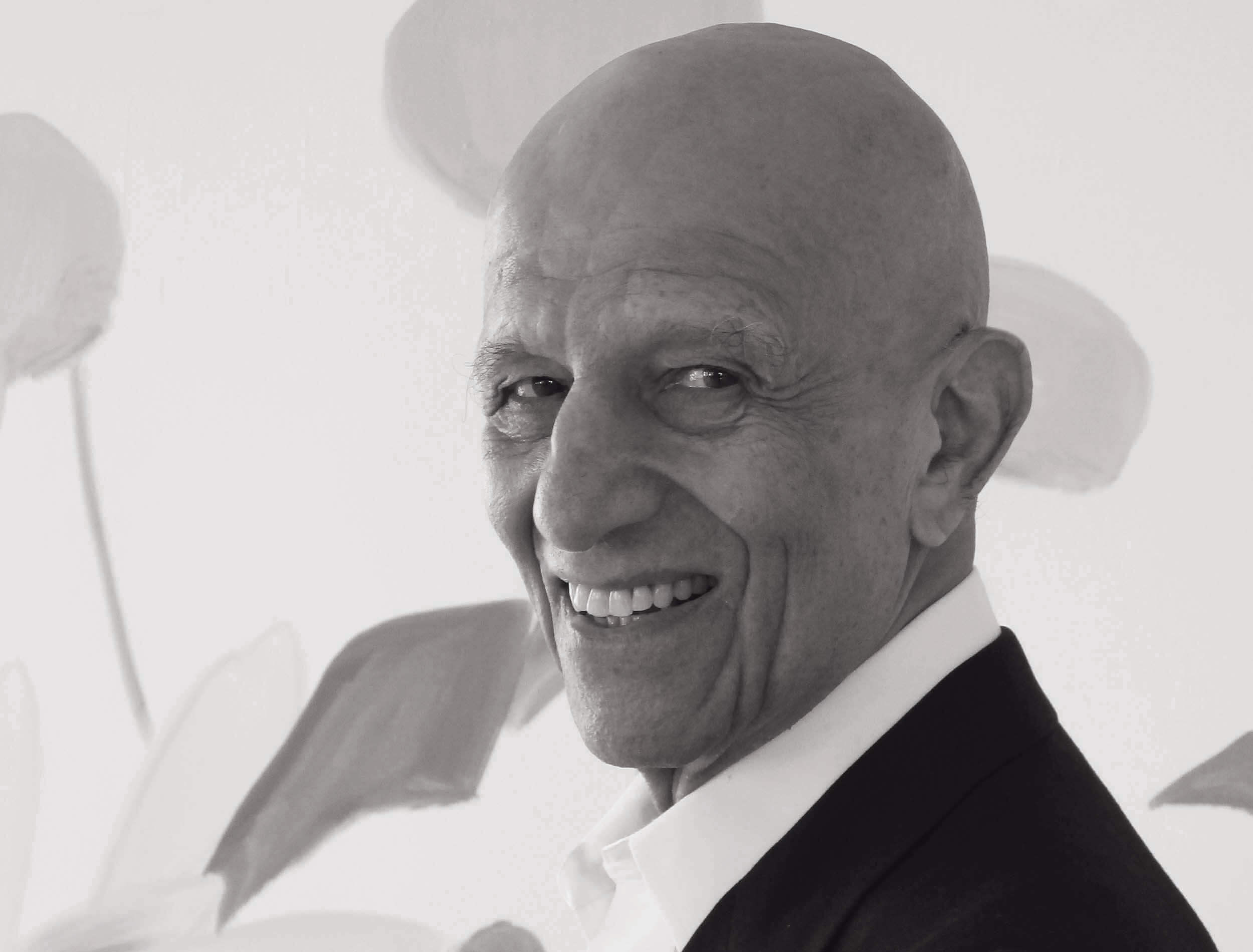 Elderly man smiling in black and white photo, wearing a suit jacket, with a floral background.
