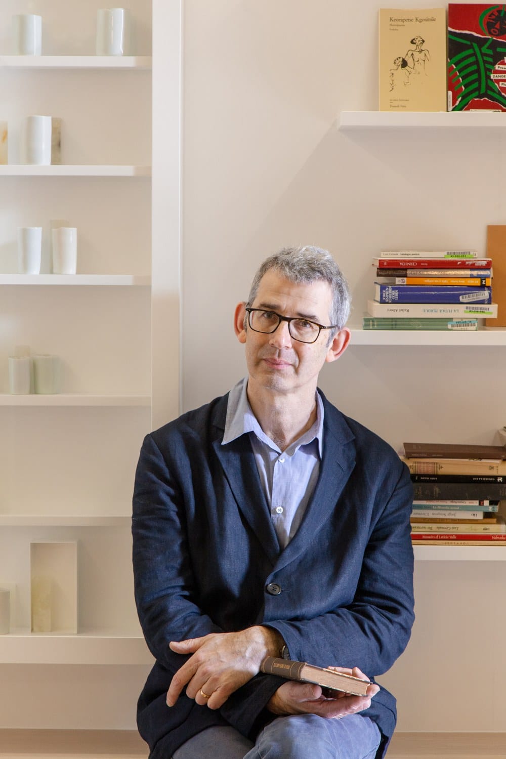 Man sitting with a book in front of shelves filled with books and vases.