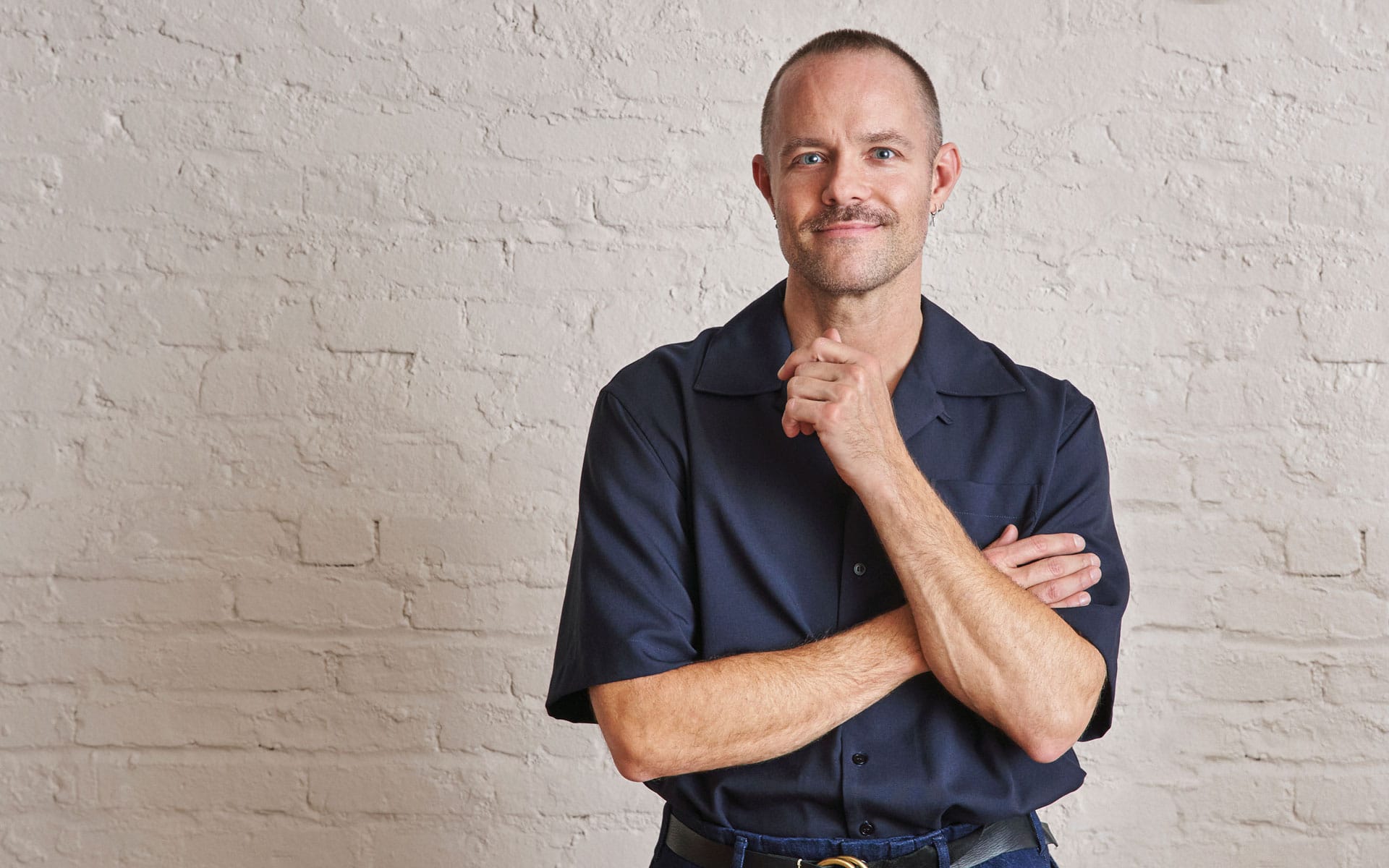 Smiling man with a mustache in a dark shirt standing against a light brick wall.