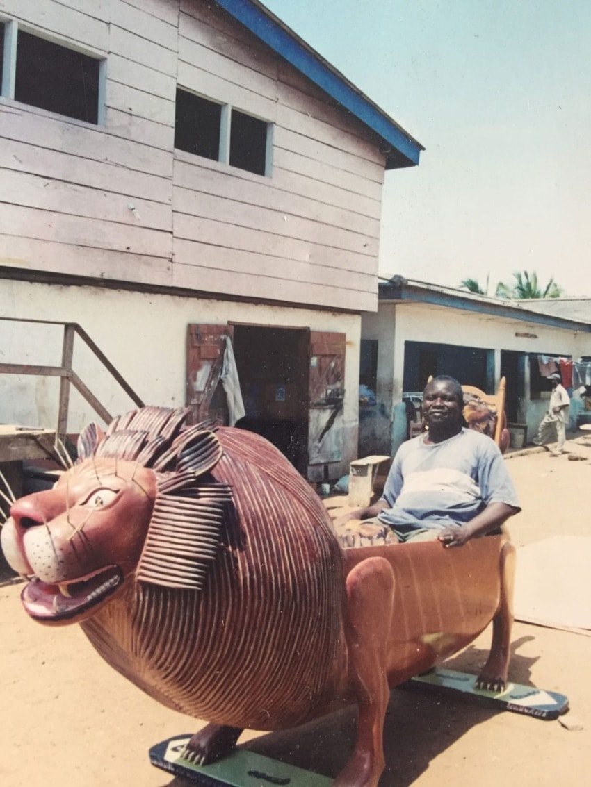 Man sitting on a carved wooden lion ride, outside a building with a wooden facade, on a sunny day.