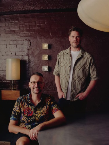 Two people in a dimly lit room with brick walls and a modern lamp, one seated and one standing, both wearing casual shirts.