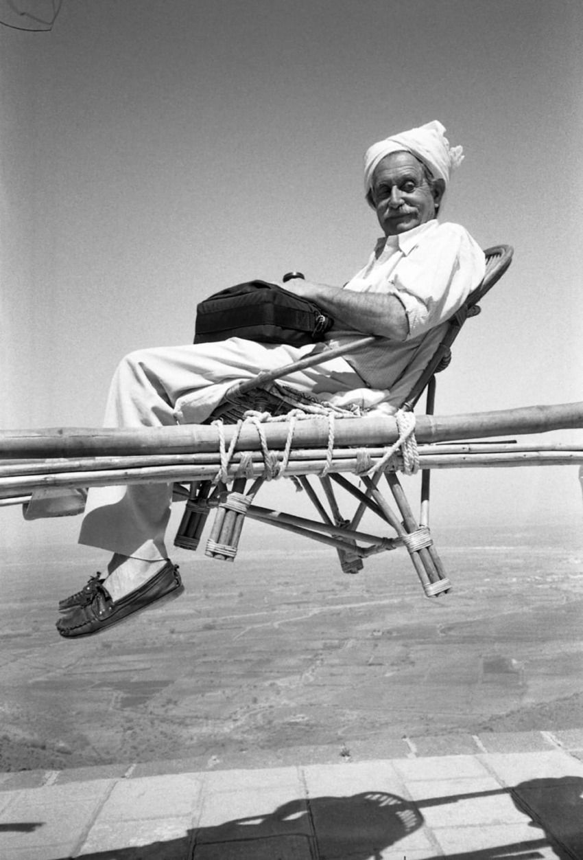 Man in traditional attire sitting on a chair high above a landscape with a serene expression and a clear sky in the background.