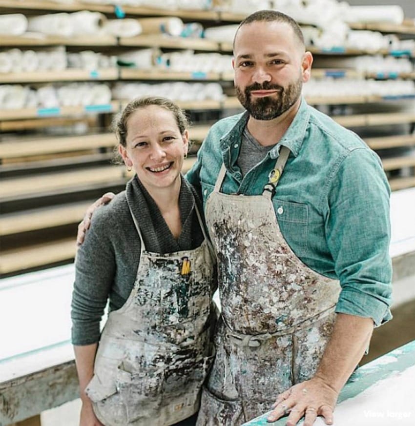 Two artists wearing paint-splattered aprons standing together in a workshop with shelves of rolled canvas behind them.