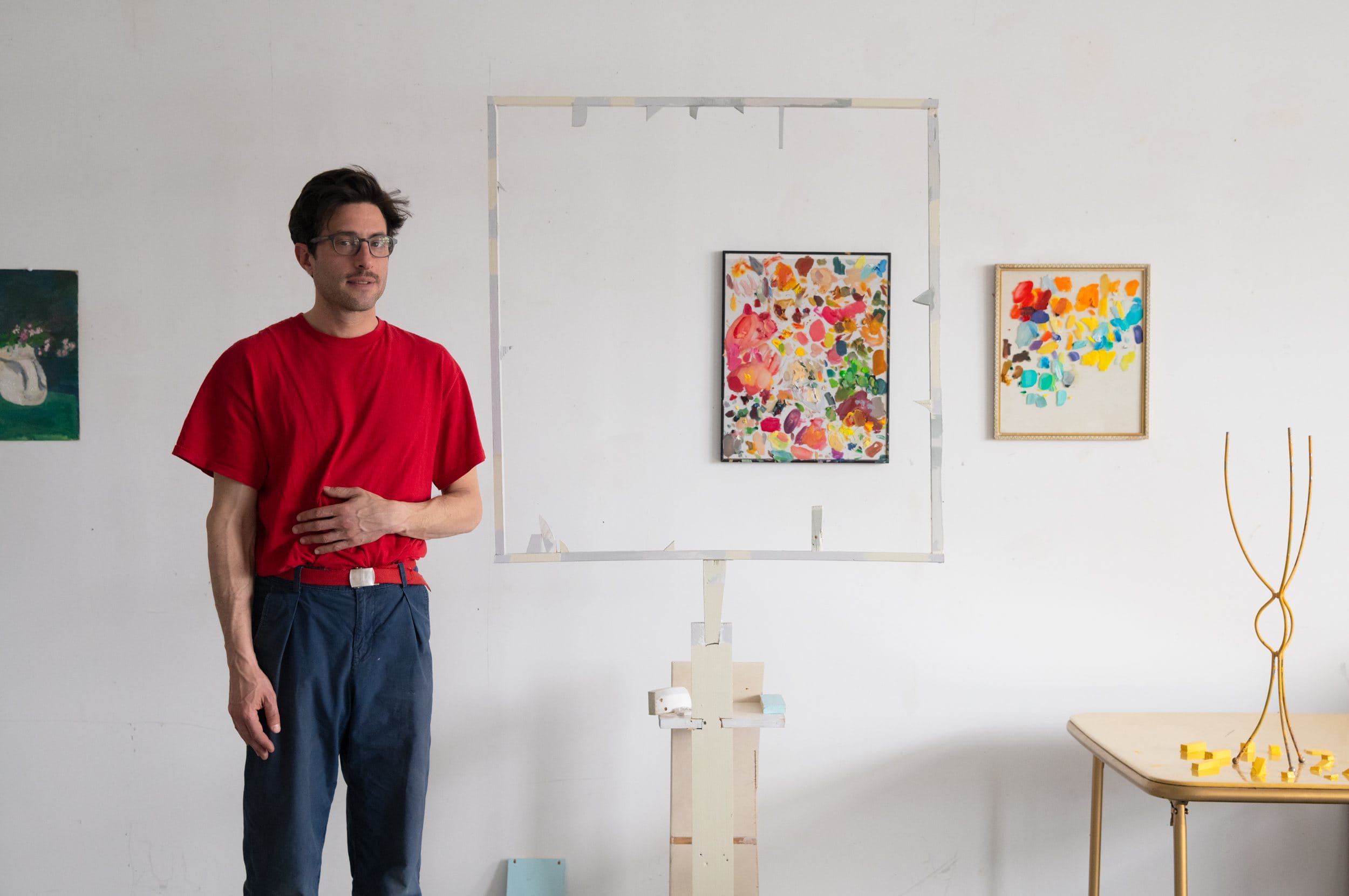 Man in red shirt standing next to colorful abstract paintings on a white wall in an art studio.