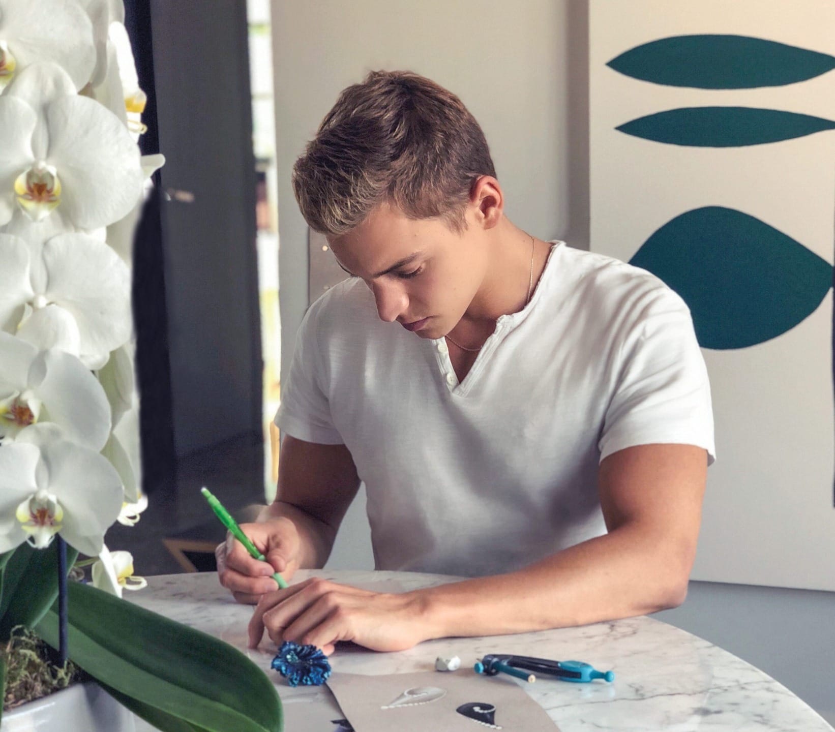 Man in white shirt writing at a table with flowers and art around him.