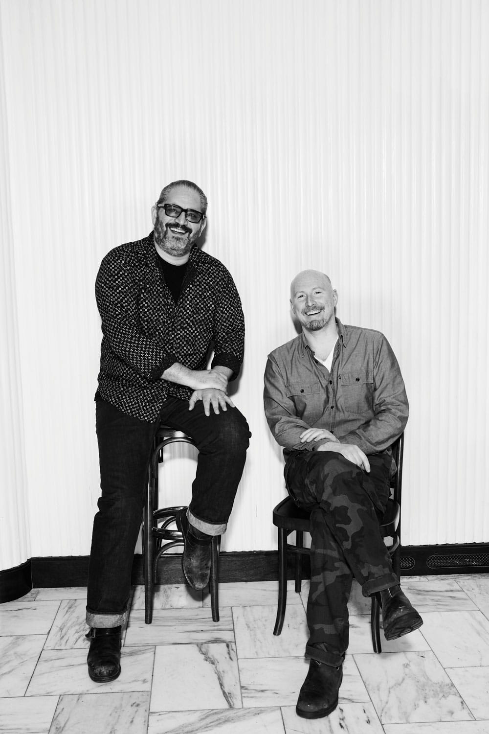 Two men sitting on chairs, one smiling and one laughing, against a white wall indoors.