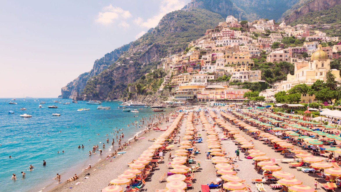 Beach with colorful umbrellas, people swimming, and a hillside town with vibrant buildings in Positano, Italy.