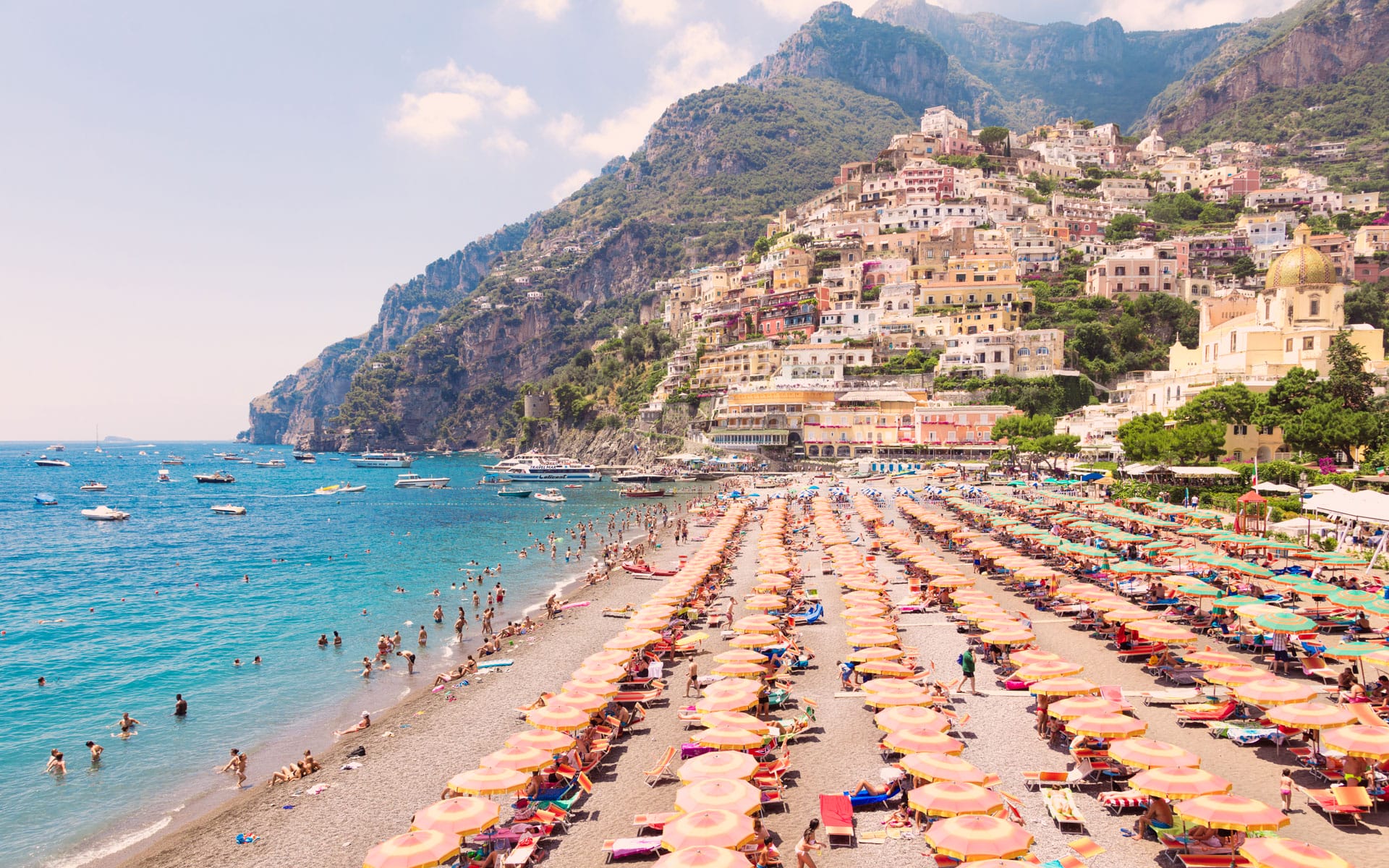 Beach with colorful umbrellas, people swimming, and a hillside town with vibrant buildings in Positano, Italy.