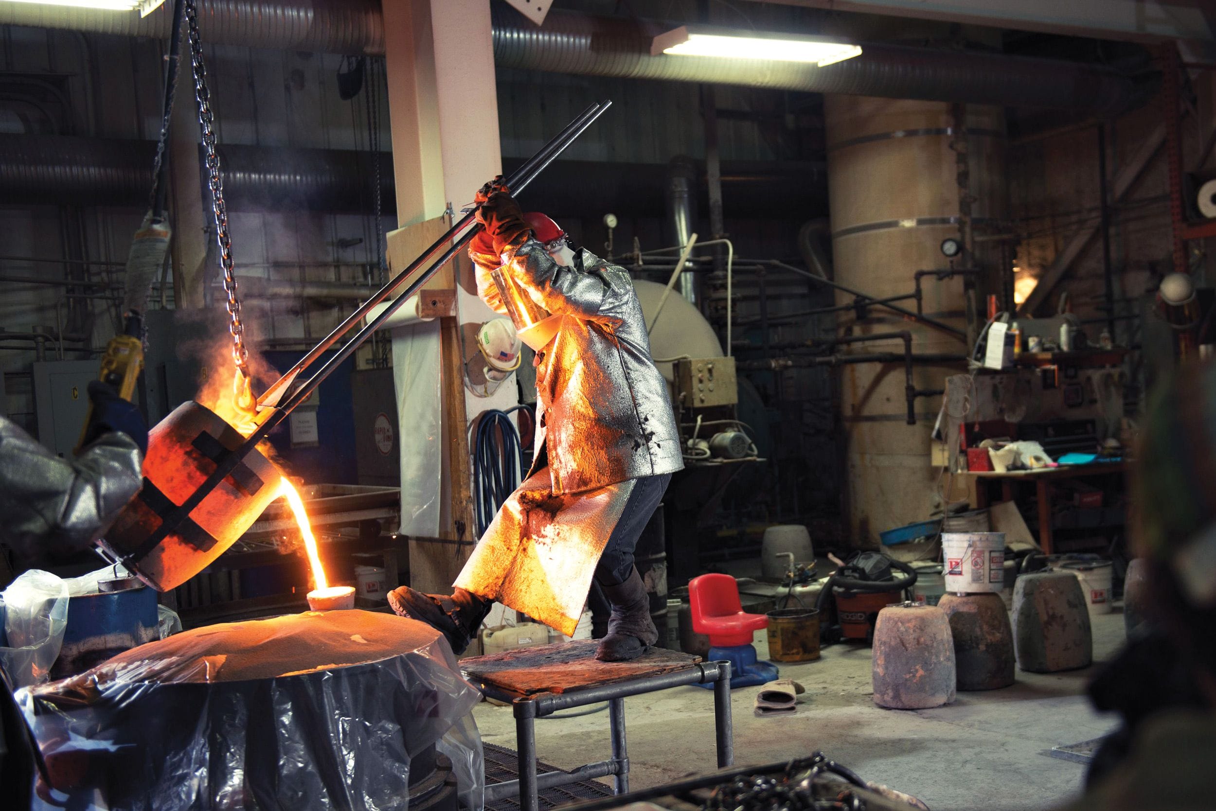 Worker pouring molten metal in foundry, wearing protective gear in industrial workshop setting.