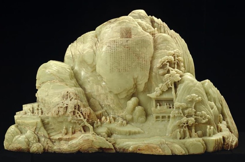 Intricately carved jade mountain sculpture featuring trees, buildings, and tiny figures against a dark background.