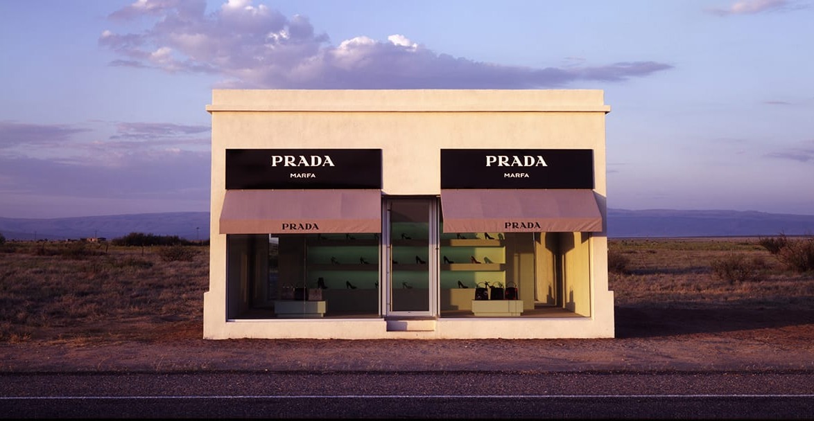 Exterior view of Prada Marfa art installation, resembling a small storefront, set in a desert landscape under a cloudy sky.