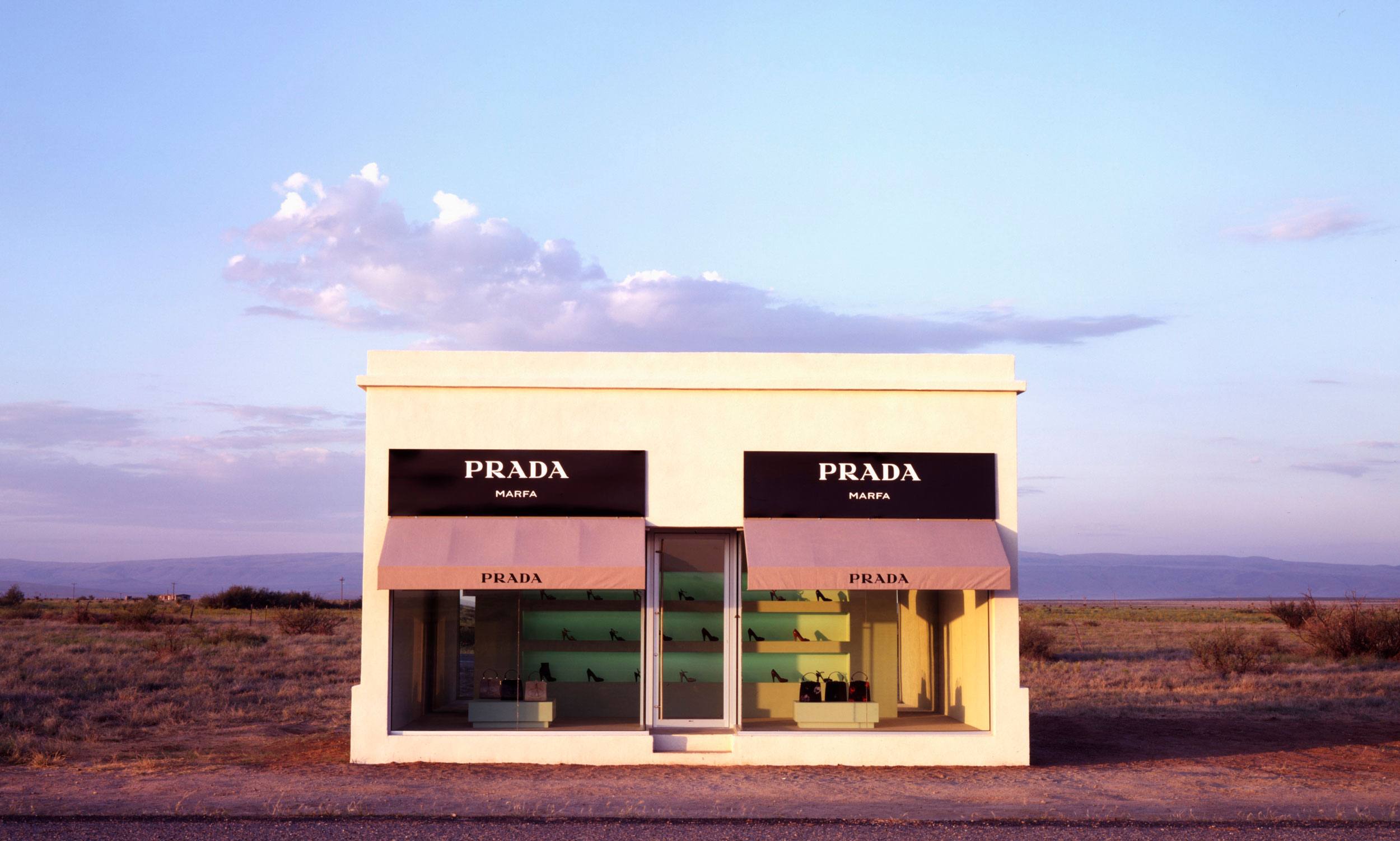 Desert landscape with a small white building featuring Prada signage, clear sky in the background, and a barren foreground.