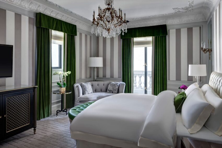 Elegant hotel room with striped wallpaper, a chandelier, green curtains, white bedding, and a small seating area near a window.