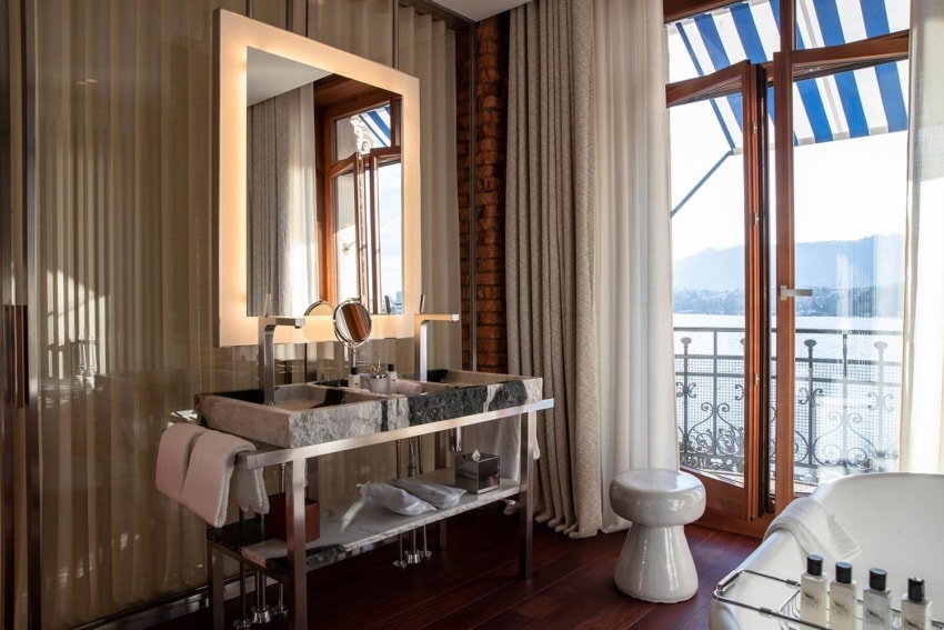 Luxurious bathroom with a marble sink, large mirror, striped curtains, and a scenic view of a lake through the balcony door.