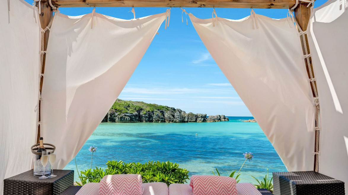 Beach cabana with a view of turquoise ocean and distant rocky cliffs, surrounded by lush greenery under clear blue skies.