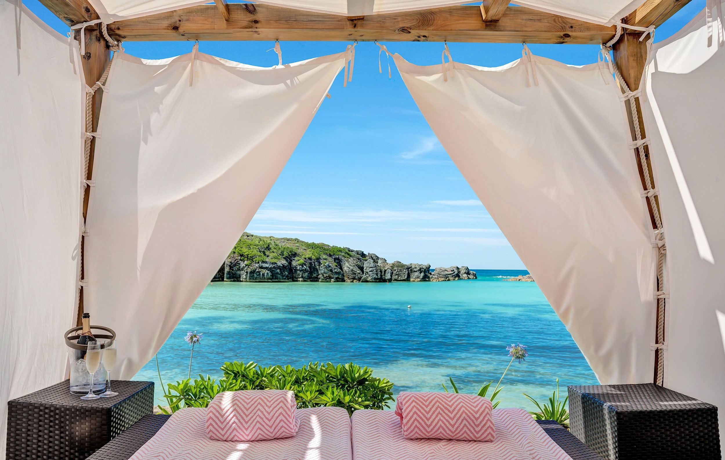 Beach cabana with a view of turquoise ocean and distant rocky cliffs, surrounded by lush greenery under clear blue skies.