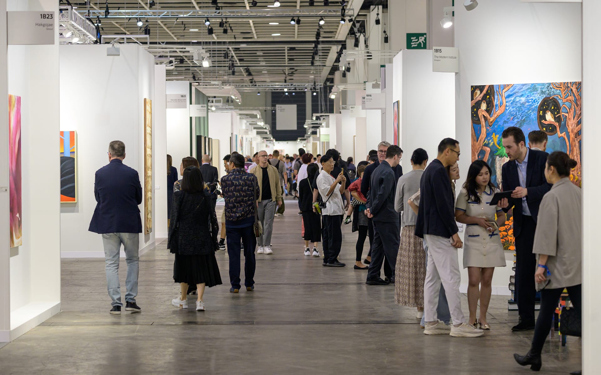 People viewing art at a modern gallery exhibition, walking through a spacious hallway with various artworks on display.