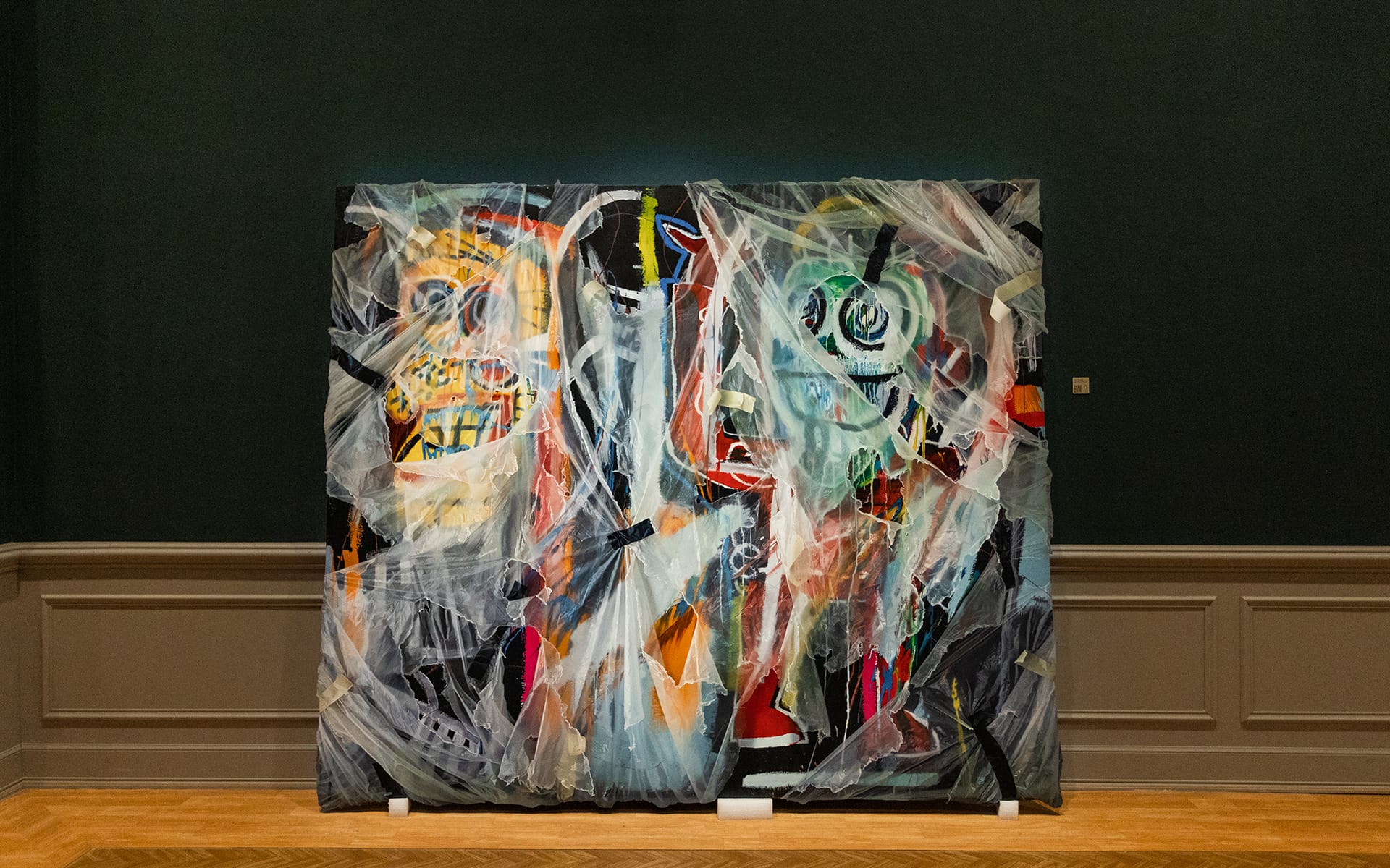 Abstract painting with vibrant colors displayed in a gallery against a dark green wall and wooden floor.