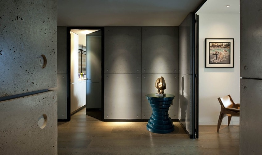 Modern interior hallway with concrete walls, wooden chair, abstract sculpture on blue pedestal, and framed artwork on wall.