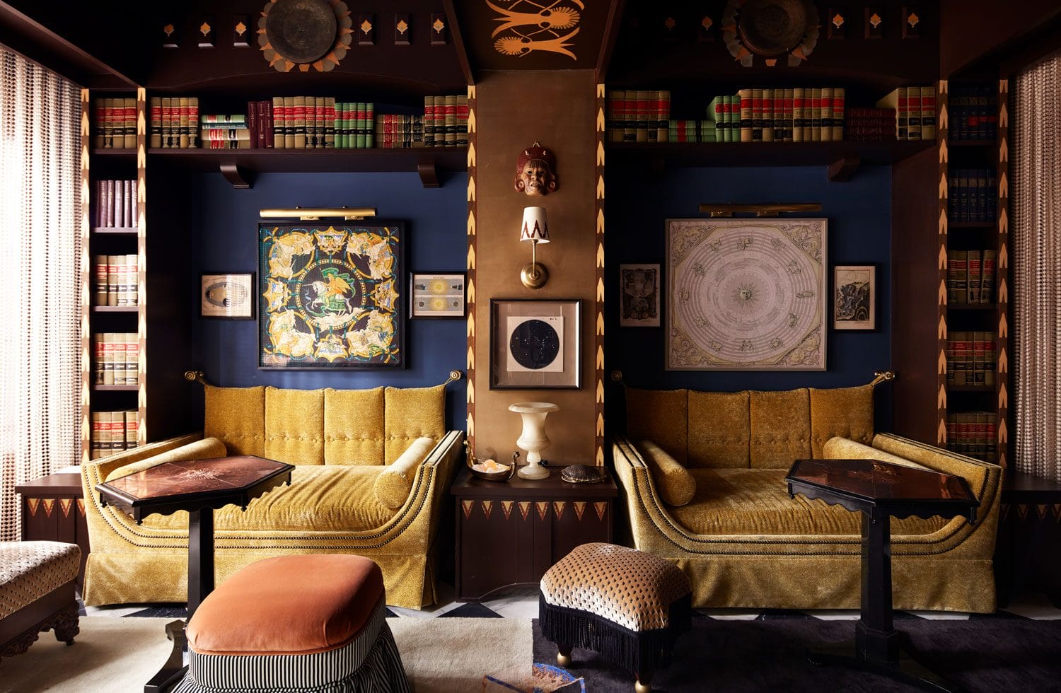 Luxurious living room with gold couches, ornate decor, art-filled walls, and bookshelves adorned with colorful books.