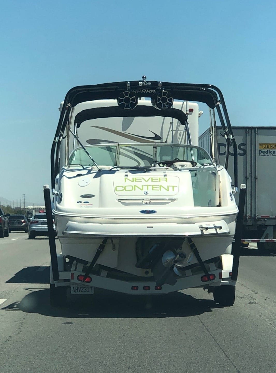 Boat towed on highway displaying the name "Never Content" on its stern, with blue sky and traffic in the background.
