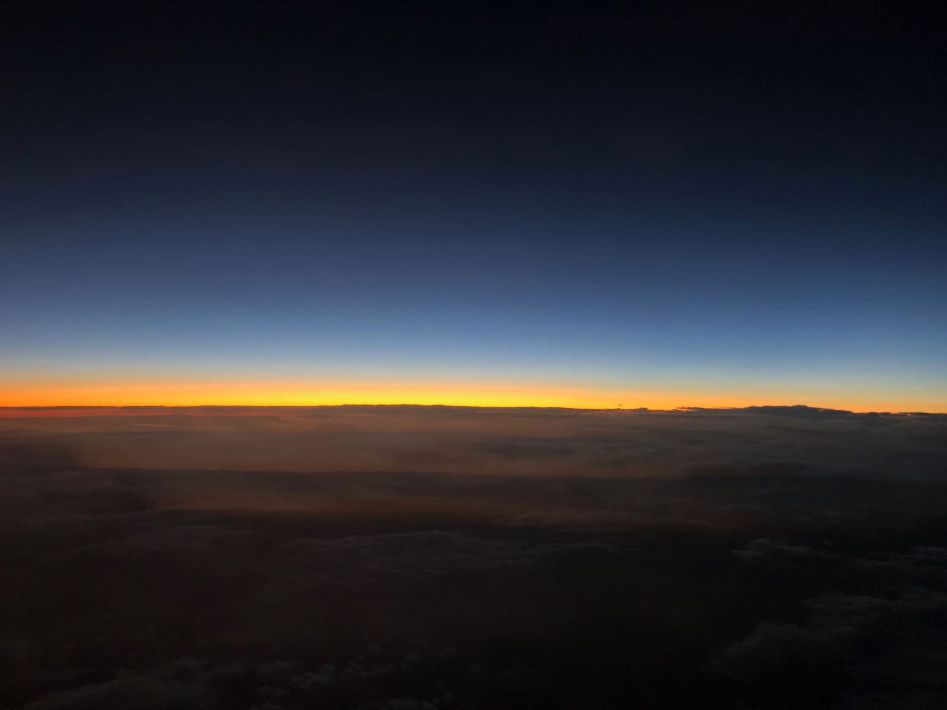 Colorful horizon at sunset seen from an airplane window, with dark clouds below and a gradient sky above.