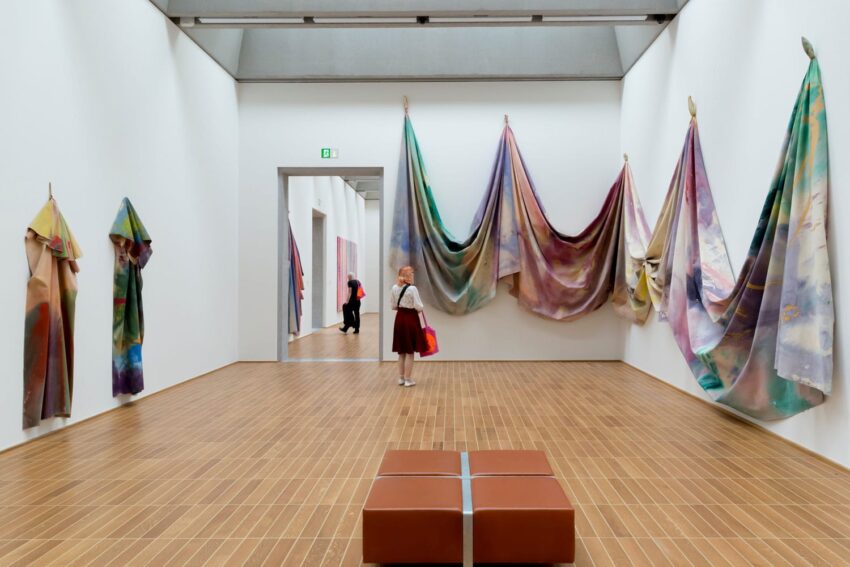 Woman observing colorful draped fabrics in a modern art gallery with wooden floors and white walls.