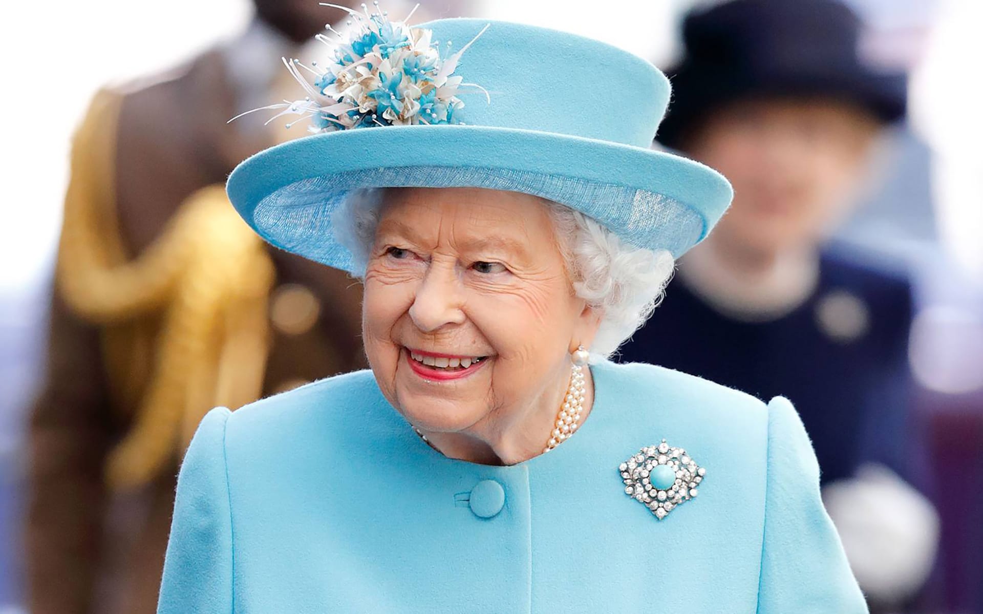 Elderly woman in blue outfit with matching hat, adorned with pearls and brooch, smiling during an outdoor event.