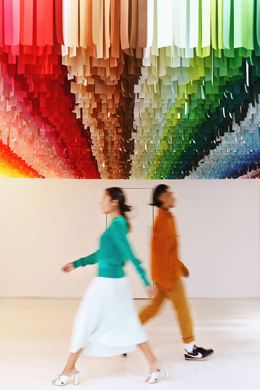 Two people walking under a vibrant, colorful installation of hanging paper strips in an art gallery.