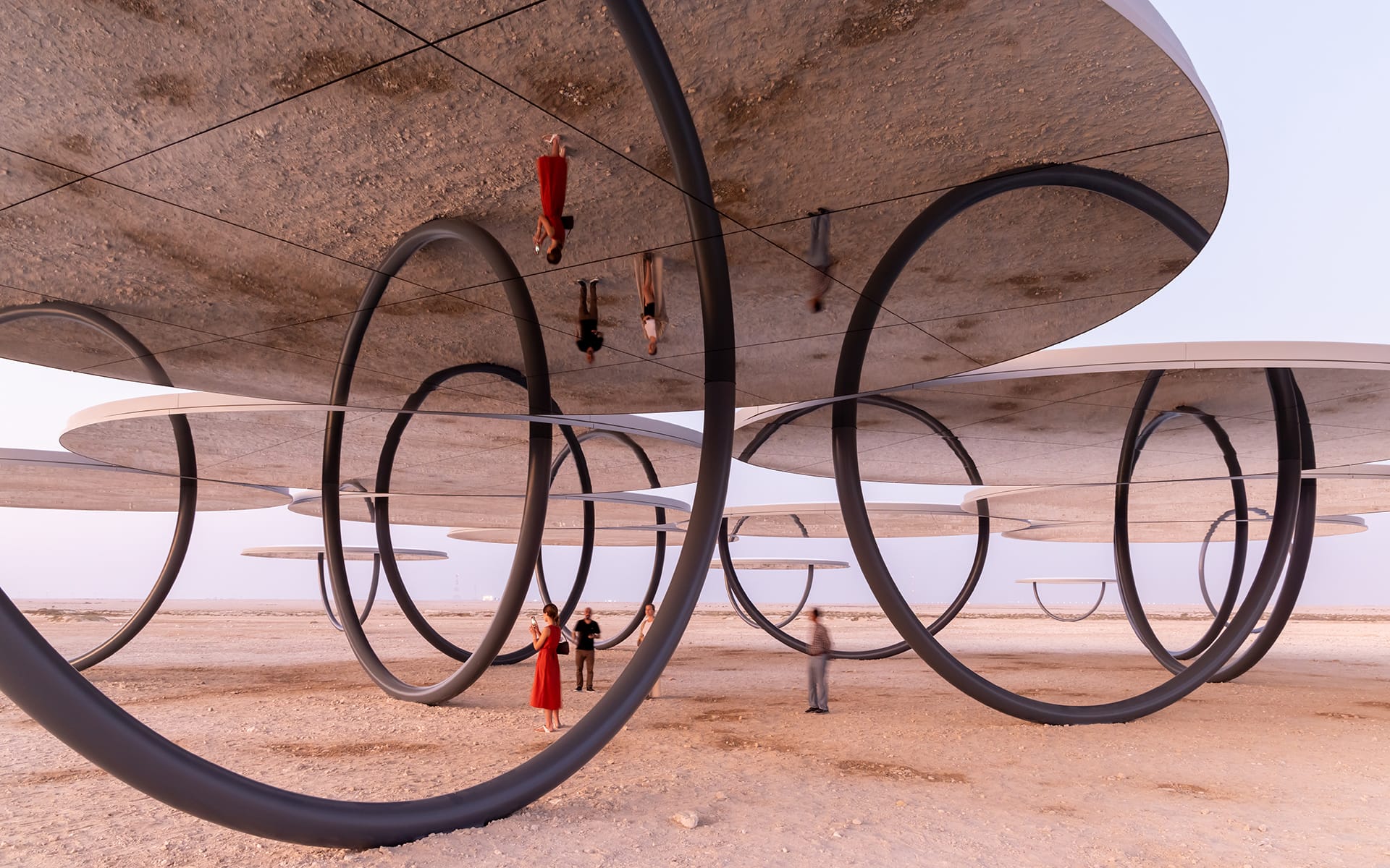 People walking and standing under large circular metal structures in a desert landscape.