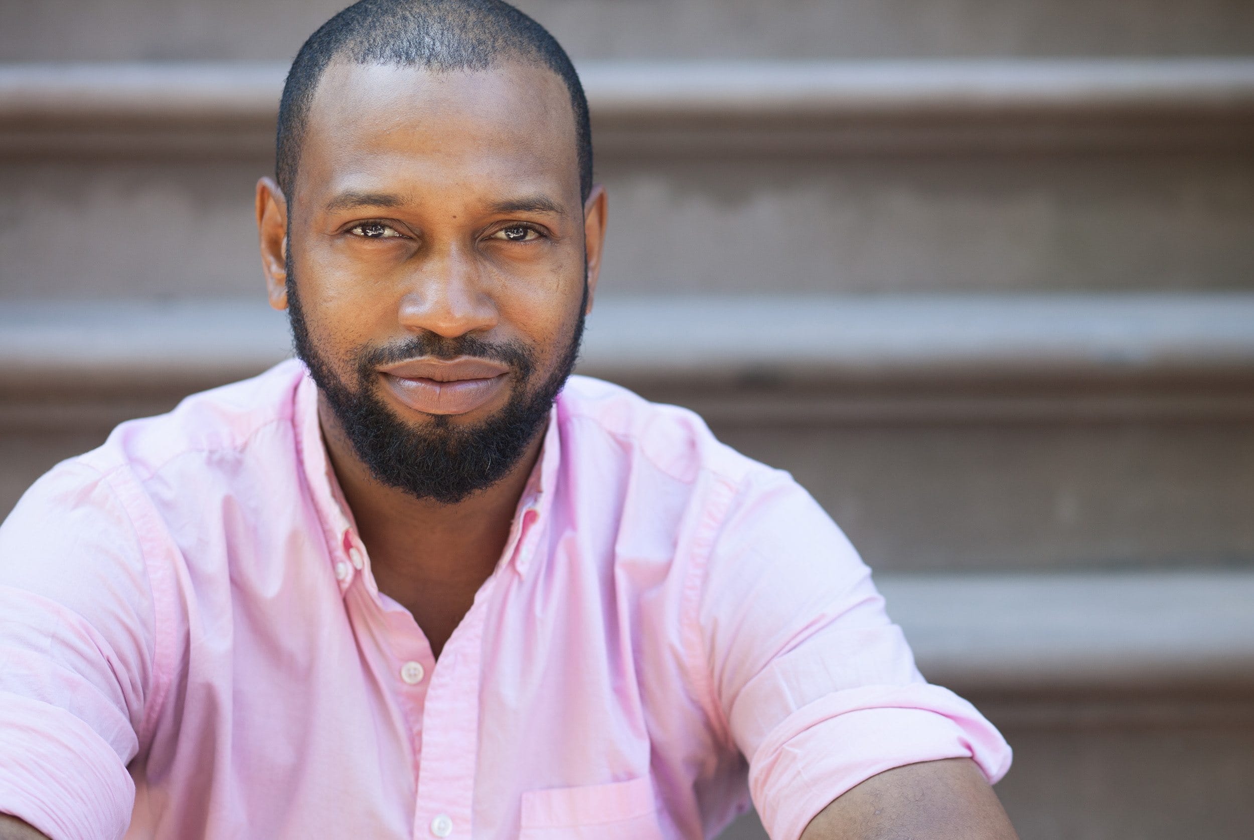 Man sitting on steps wearing a pink shirt, looking directly at the camera with a slight smile.