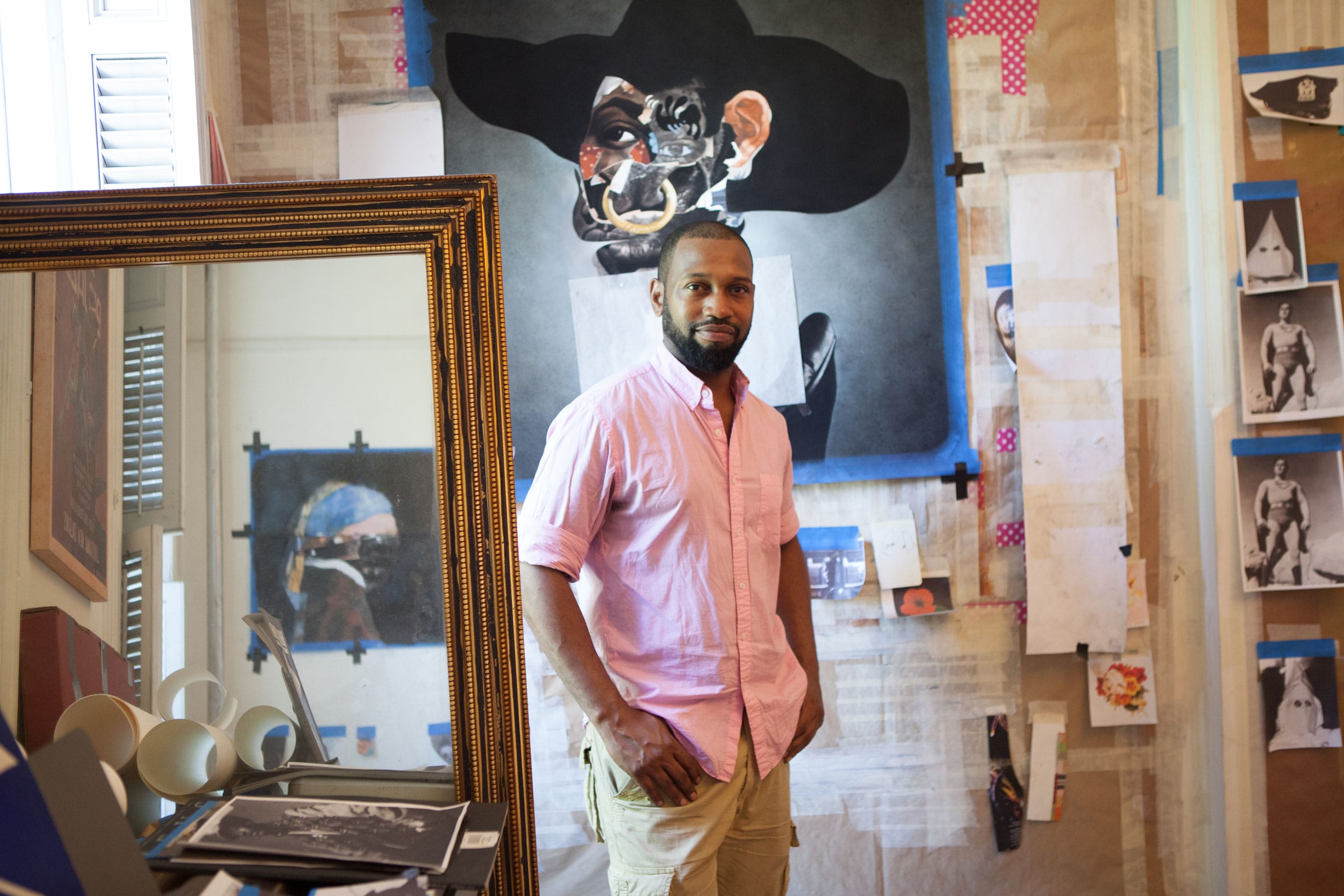 Artist in a studio with colorful artwork and various sketches on the walls, wearing a pink shirt and beige pants.