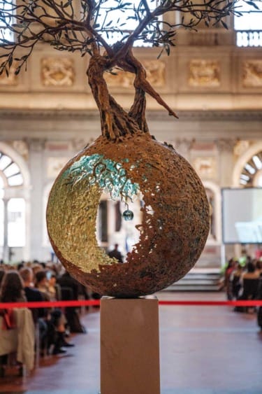 Sculpture of a tree growing from a hollow, textured sphere in an ornate building interior.