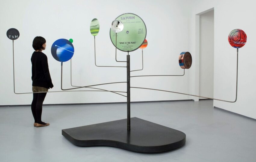 Woman observing a modern art sculpture featuring various colored discs on metal rods in a gallery setting.