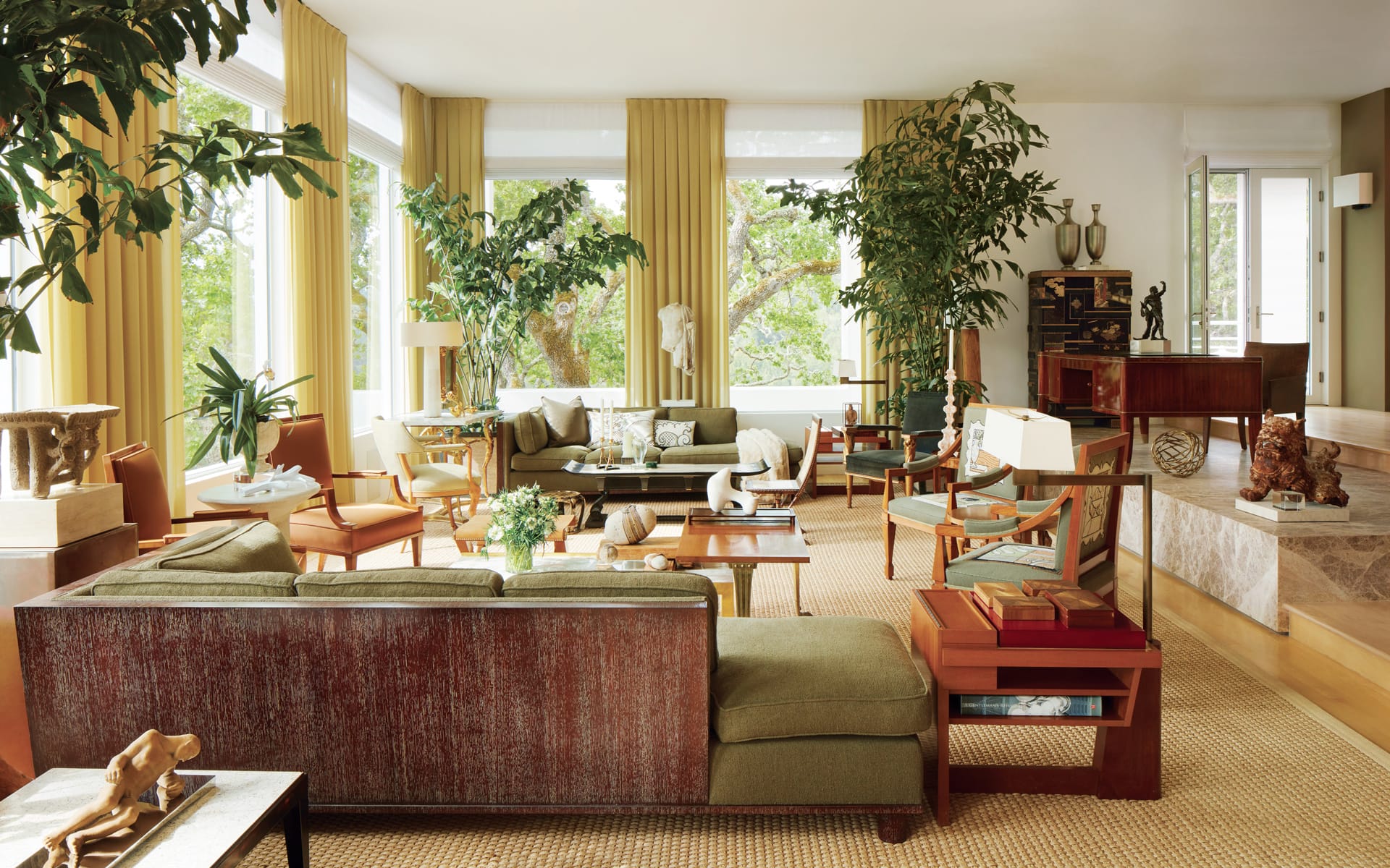 Spacious living room with green sofas, large windows, indoor plants, and wooden furniture.