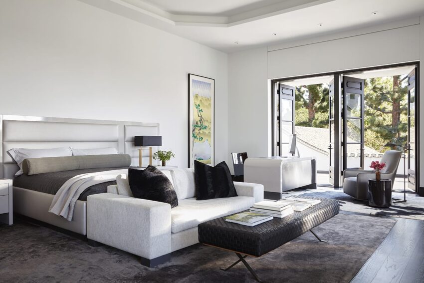 Modern bedroom with large bed, plush pillows, art on walls, glass doors opening to greenery, and a cozy seating area.