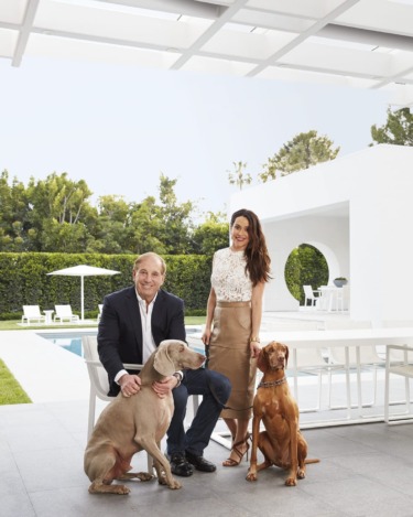 Couple smiling with two dogs by their side, seated outdoors on a modern patio with a pool and greenery in the background.