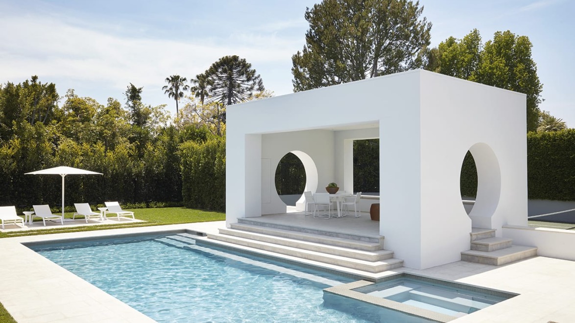 Modern backyard with a white pergola, swimming pool, lounge chairs, and greenery on a sunny day.