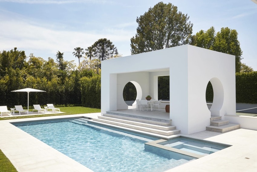 Modern backyard with a white pergola, swimming pool, lounge chairs, and greenery on a sunny day.