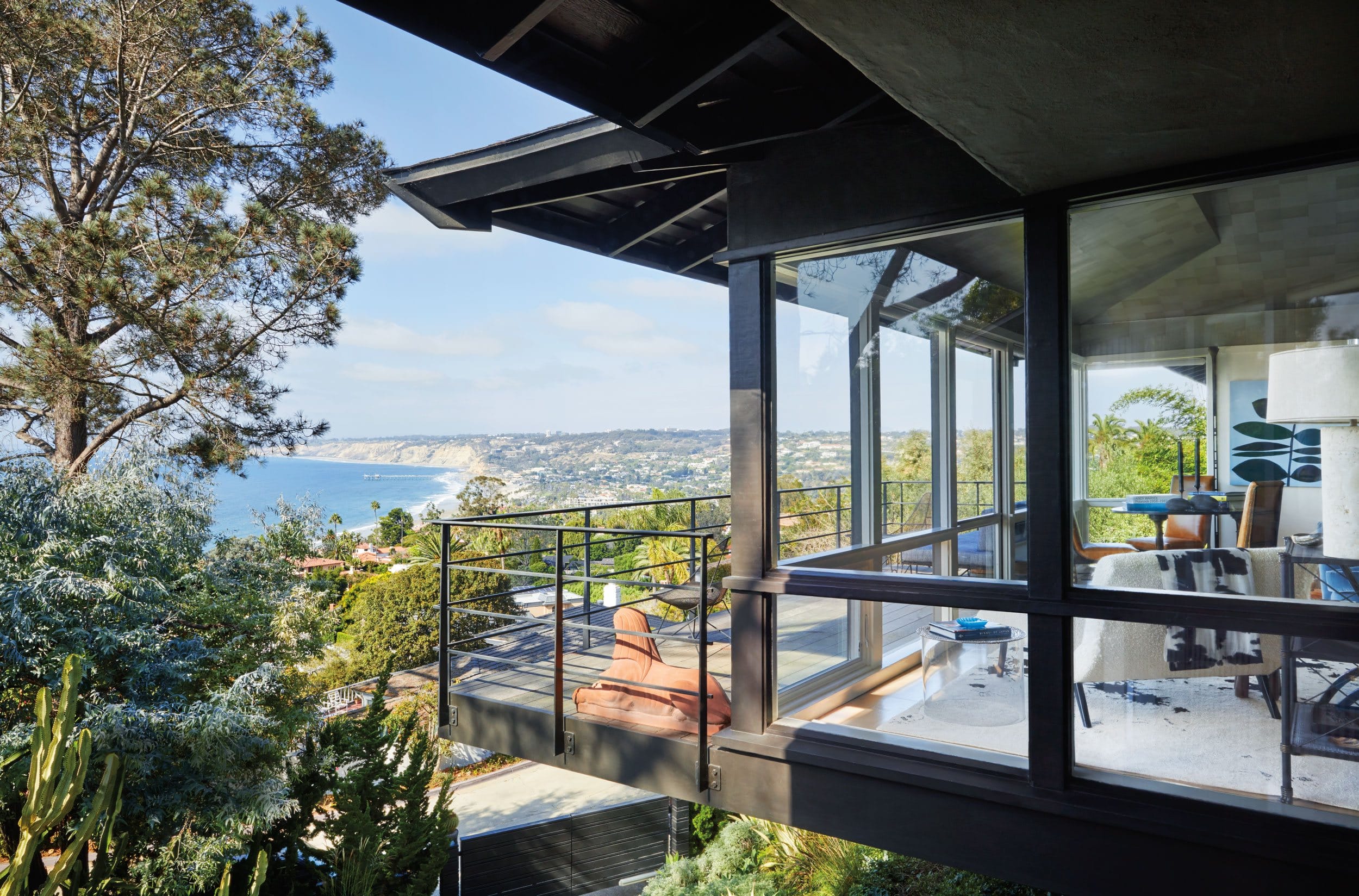 Modern house with glass walls offering ocean and hillside views, surrounded by trees and greenery.